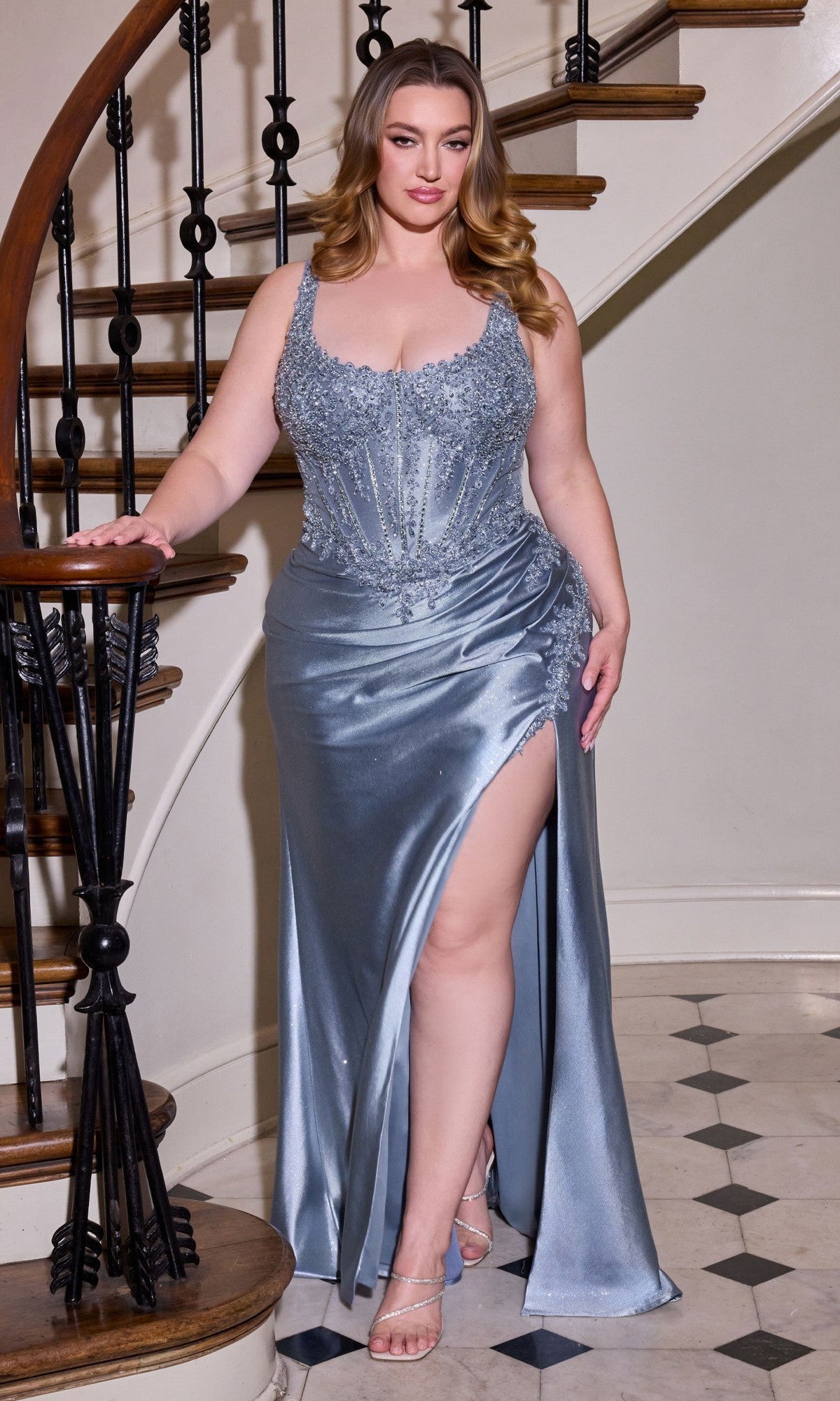 A woman stands on a staircase in the Ladivine CC475C, a silver-blue satin long plus-size prom dress featuring a corset bodice, thigh-high slit, and beaded details, her hand resting on the banister.